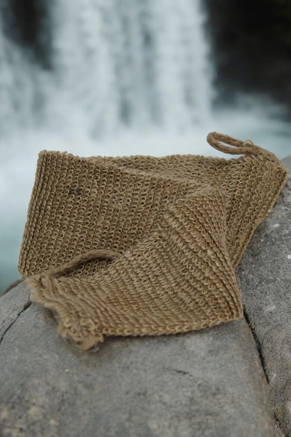 An All Natural Back Scrubber from The Lather and The Wick is casually draped over a large rock in the foreground. In the background, a waterfall cascades down, creating a serene and natural scene. The focus is on the texture of the biodegradable back scrubber against the smooth rock with the blurred waterfall behind it.