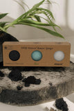 A box of The Lather and The Wick's Konjac Sponge - Facial Exfoliator 3 pack features three varieties: Bamboo/Charcoal (black), Aloe (green), and Natural (white). The box is beautifully displayed on a textured stone surface with lush green leaves in the background.