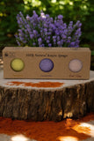 A box of Konjac Sponge - Facial Exfoliator 3 pack from The Lather and The Wick, labeled as Turmeric, Lavender, and Red Clay stands on a tree stump. Purple lavender flowers bloom in the background, while a spread of orange turmeric powder is visible on the stump—a perfect blend for a rejuvenating facial exfoliator.