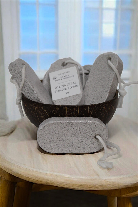 A wooden bowl filled with rectangular, gray pumice stones from The Lather and The Wick, each attached to a white rope loop. One pumice stone has a tag that reads 