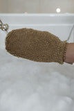 A hand wearing The Lather and The Wick's All Natural Bath Mitt hovers over a bathtub filled with bubbles, ready to exfoliate. The background reveals a bathroom adorned with white tiles, highlighting the mitt's biodegradable nature.