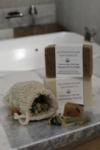 A stack of two soaps labeled "Dragon's Fire" and "Lavender Fields" from The Lather and The Wick company is placed near a bathtub. In front of the soaps is a Pre-filled All Natural Soap Saver with dried herbs and botanicals spilling out.